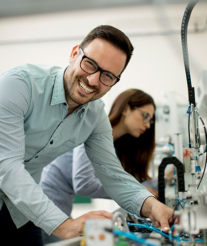 Hombre con gafas y camisa a rayas sonriendo, trabajando con maquinaria junto a una mujer con gafas de protección